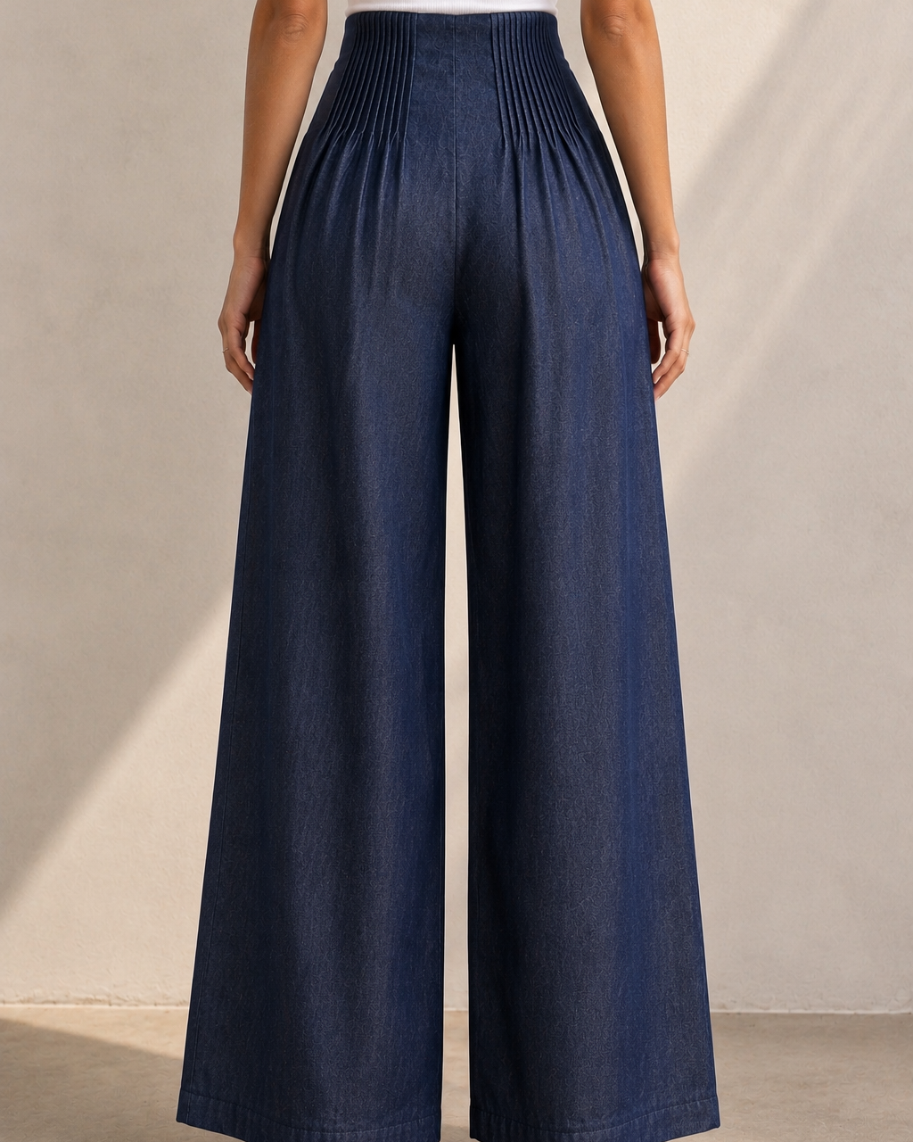 A model shown from the back wearing high-waisted, wide-leg pleated pants in a dark blue denim-like fabric, paired with a white top and gold heels, against a neutral beige wall.