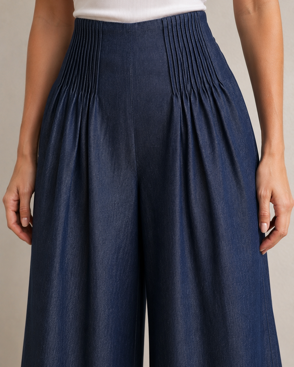 A close-up of a model wearing high-waisted, wide-leg trousers in a dark navy blue fabric with pleated detailing at the waist, paired with a white ribbed top.
