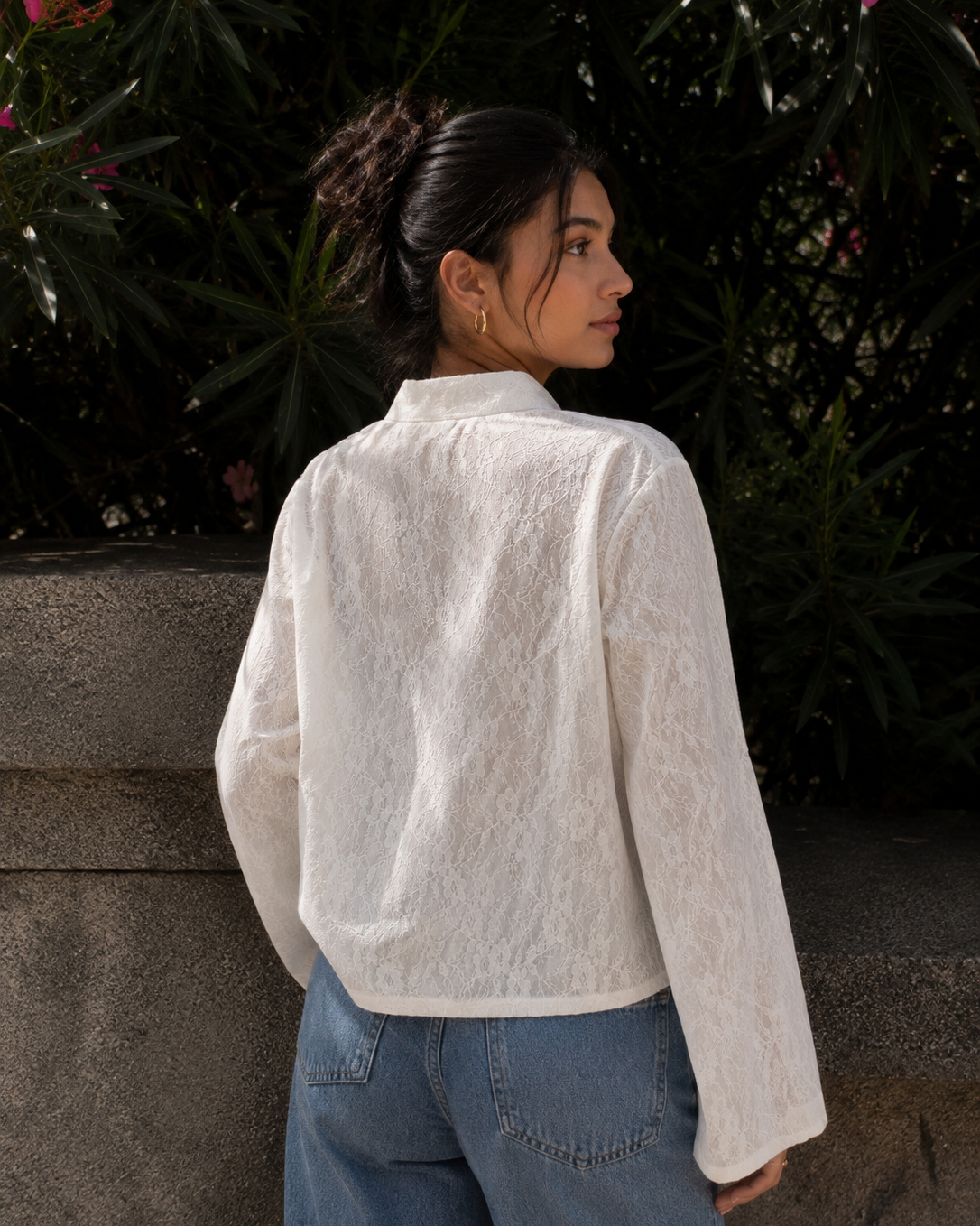 A woman wearing a white textured lace long-sleeve blouse and blue jeans, photographed from the back in an outdoor setting with dark green foliage and a stone wall behind her.
