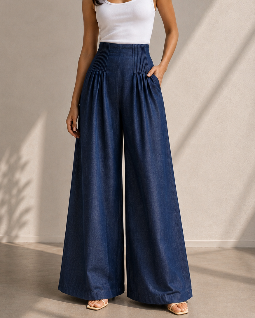 A woman modeling a pair of high-waisted, wide-leg blue pants with a white tank top, standing against a textured beige wall with soft shadows.