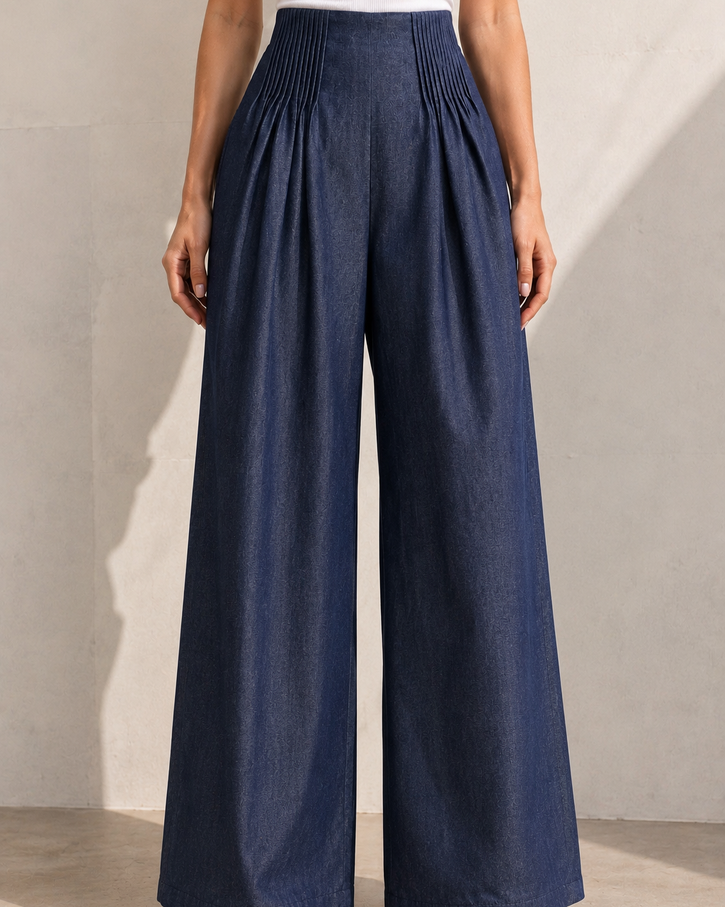A model wearing high-waisted, wide-leg pleated pants in a dark blue denim-like fabric, styled with a white top and beige sandals against a neutral concrete wall.
