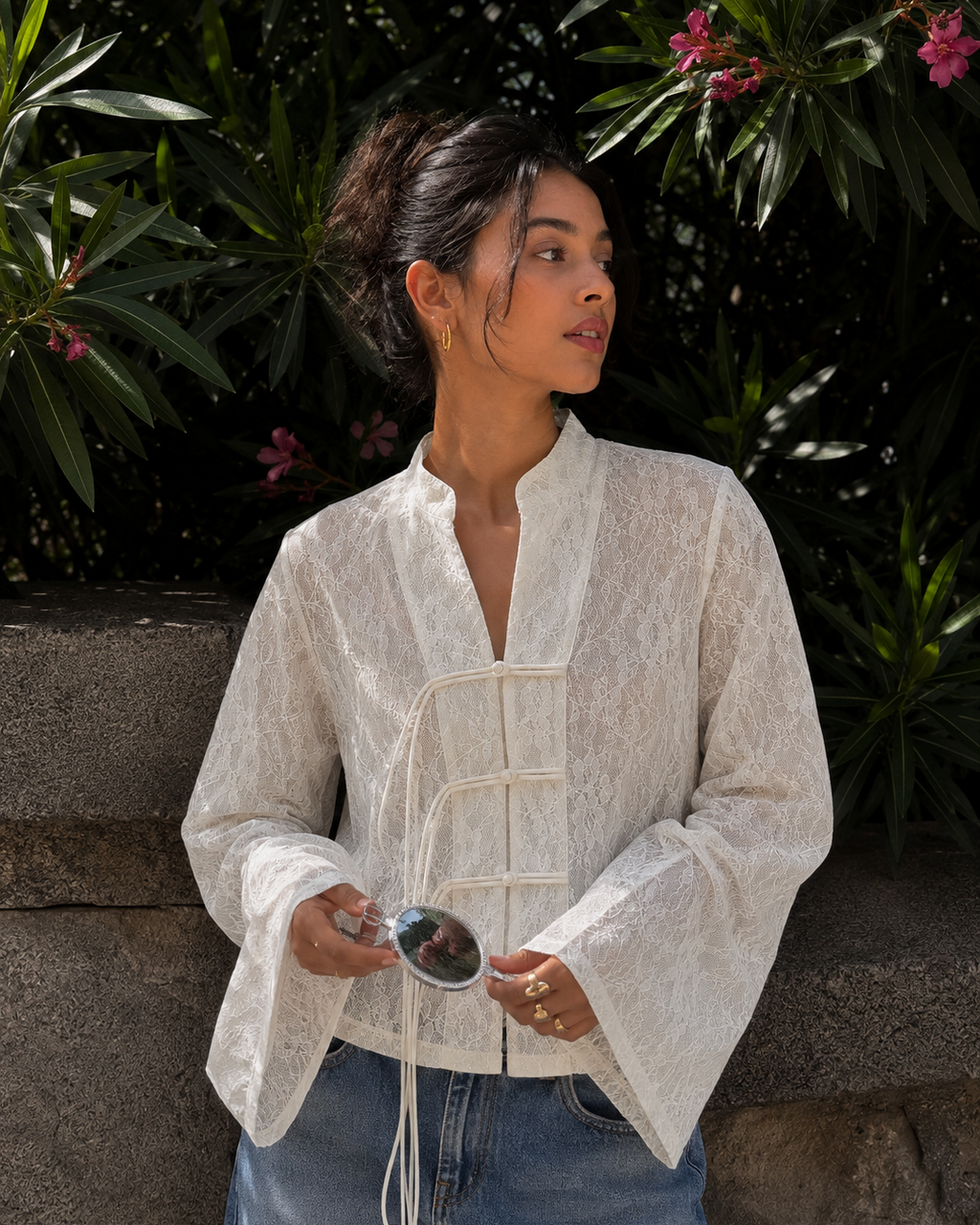 A woman wearing a white textured lace long-sleeve blouse with a mandarin collar and front tie closure, paired with blue jeans, standing outdoors in front of a stone wall and green foliage.