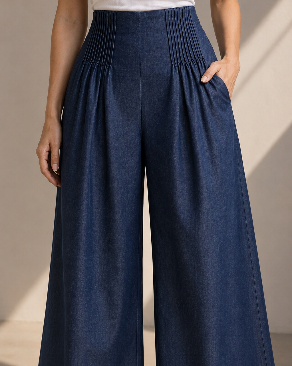 A model wearing high-waisted, wide-leg pleated trousers in a dark navy blue color, paired with a white sleeveless top. The photo is taken in a warm, sunlit indoor setting with soft shadows.