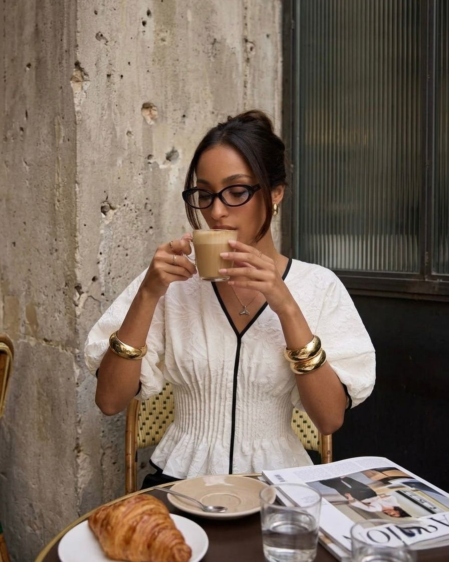 Woman drinking coffee and reading a magazine at an outdoor cafe.